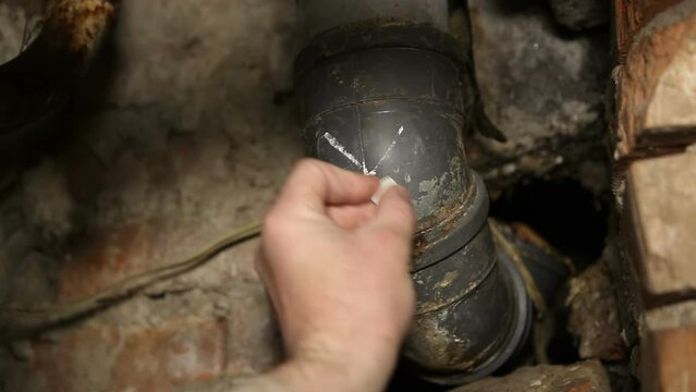 A male plumber marks a plastic sewer pipe with a cross for repair and replacement. Old sewer pipes in the basement of a house.