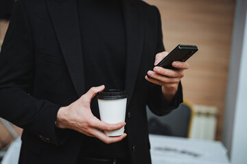 Man in black suit holds coffee and cell phone, making a call