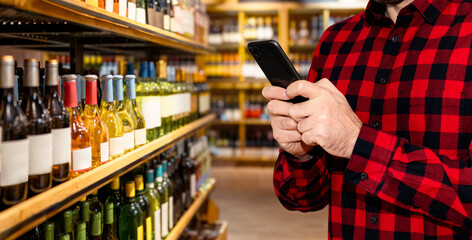 Male retail shopper uses his smartphone and the Internet while choosing wine in a liquor store.