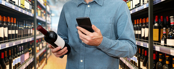 Male retail customer uses his mobile phone and the Internet while choosing wine in a liquor store.