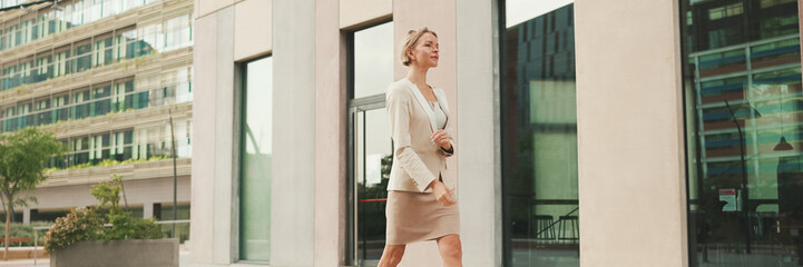 Businesswoman in beige suit walks outside past business building, Panorama