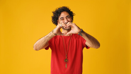 Fototapeta na wymiar Happy attractive man with curly hair wearing red T-shirt, showing representing heart in shape of fingers gesture looking at camera isolated on yellow background in studio