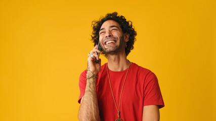 Smiling man with curly hair, dressed in red T-shirt,  talking on mobile phone isolated on yellow...