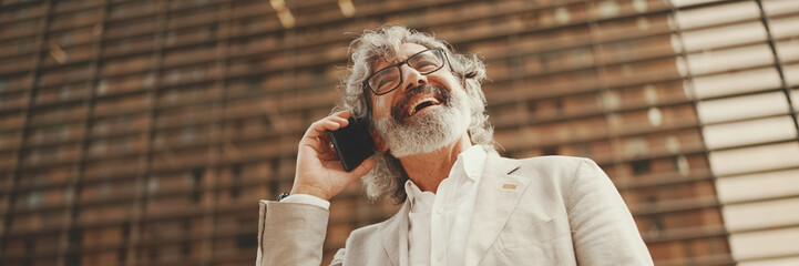 Clouse up, mature businessman with beard in eyeglasses wearing gray jacket is using cell phone, Panorama. Middle aged manager talking on smartphone while sitting outside office - Powered by Adobe