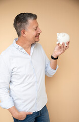 mid age man with grey hair with his white piggy bank in front of brown background, money, hammer