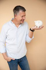 mid age man with grey hair with his white piggy bank in front of brown background, money, hammer