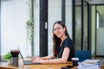Cheerful young woman working on her laptop at a wooden table in a bright, plant-filled outdoor setting.