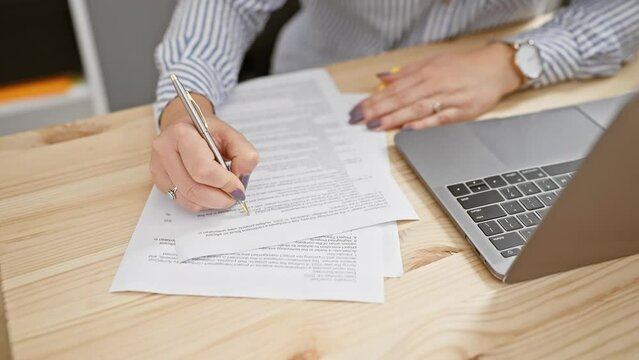 A focused woman in glasses with long hair working on documents at a modern office desk with a laptop.