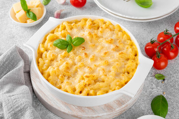 Mac and cheese in a white baking dish on a wooden board on a gray concrete background. American cuisine, comfort food. Copy space.