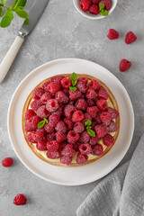 Delicious raspberry tart with vanilla custard cream sprinkled with powdered sugar on a white plate on a gray concrete background. Summer dessert.