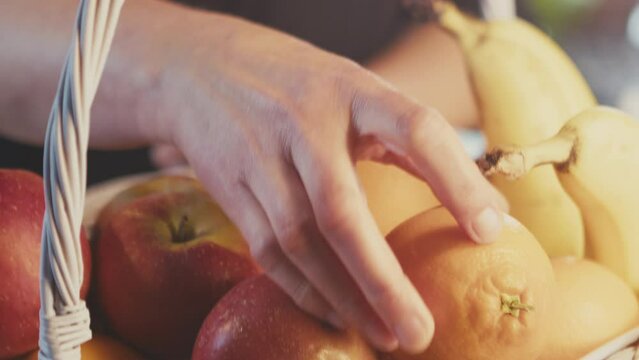 Woman's hands arranging fruits in a basket basket. Mixed fruits oranges, apples, bananas. Kitchen interior. Indoors. Studio shot lighting. Handheld. Defocused background. Black shirt. 4k footage