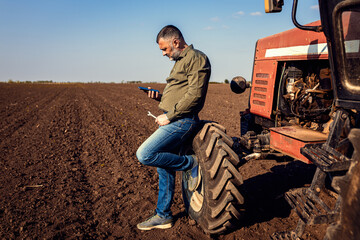 Farmer using smart phone while resting after cultivation of soil leaning on tractor tire.