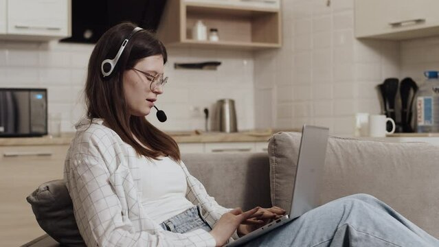 Young caucasian woman in a headset sitting on a couch at home with a laptop on her knees and talking with a client during a support service session