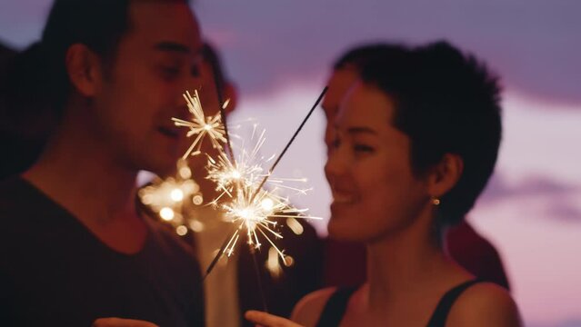 Young man and young woman standing with sparklers and cheerfully talking close up during an outdoor party with a spectacular dusk sky as a backdrop. Concept of carefree summer fun