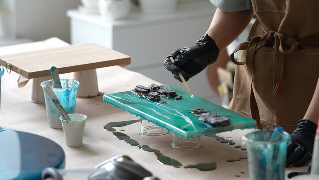 Woman artist pours epoxy resin and smears it with stick on wooden board. Craftswoman creating picture of sea foam and waves, rocks closeup view. Art deco, handmade, master class on resin art. 
