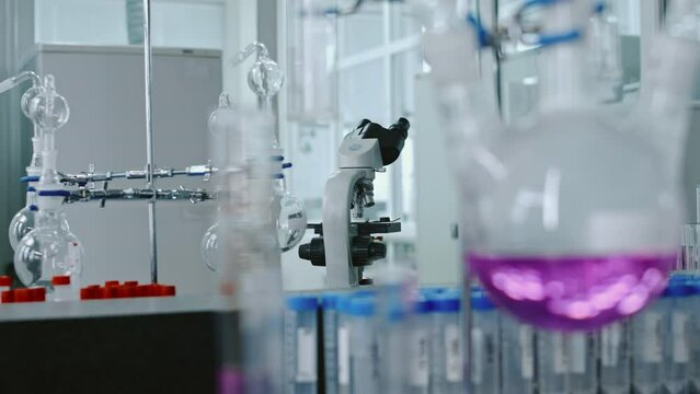 Close-up rack focus no people shot of glass retort with purple chemical fluid on stand in chemistry laboratory, rows of test tubes, microscope in backgroundClose-up rack focus no people shot of glass 
