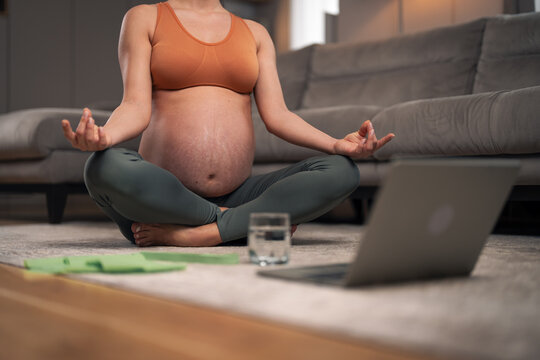 An unrecognizable pregnant woman meditating and sitting on the ground in the living room