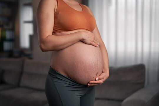 A close-up shot of an unrecognizable pregnant lady holding and caressing her belly in the living room