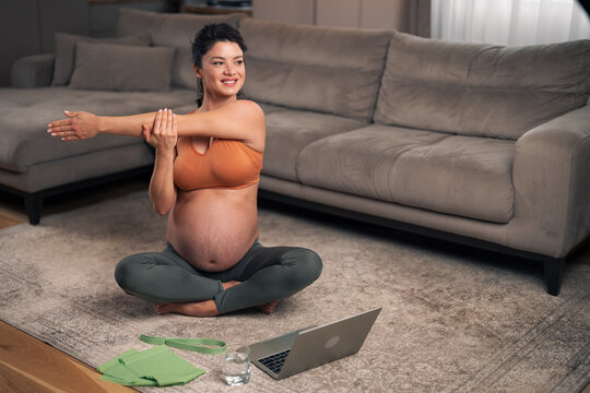 A happy adult pregnant woman stretching her arm and sitting on the ground in the living room