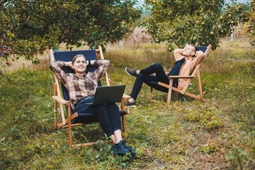 Concentrated woman in the garden working on a laptop while sitting on a chair among the trees. A woman works remotely from the garden, thinking about a new business or creative idea.