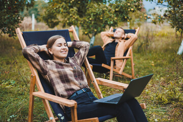 Concentrated woman in the garden working on a laptop while sitting on a chair among the trees. A woman works remotely from the garden, thinking about a new business or creative idea.
