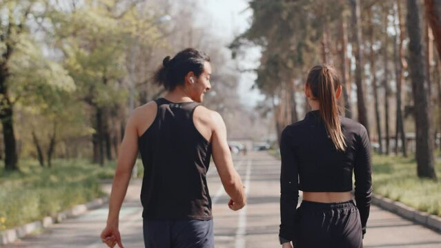 Young male athlete and young female athlete walking on an alley in an autumn city park and talking, and then start to run together during outdoor workout, back view