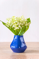 Happy May Day greeting card; bouquet of lilies of the valley in blue vase on a wooden table and white background; copy space; vertical picture