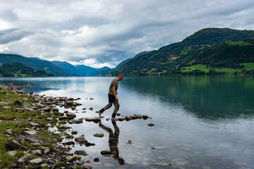 Young male traveler on the shore of mountain lake in Norway. Sports guy watching and meditating on reflections in calm pure water lake. Amazing landscape of high green mountains in Scandinavia.