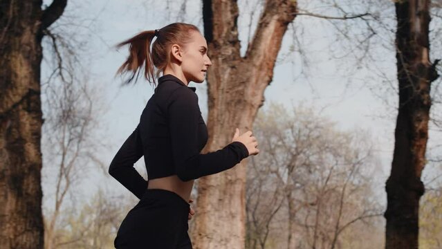 Young caucasian female athlete running leisurely on an alley in an autumn city park, side view. Concept of modern outdoor sports training in a casual urban life