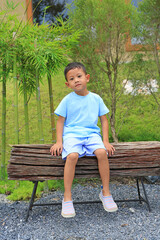 Smiling Asian little boy child looking at camera while sit on wooden chair in the garden. - 793793862