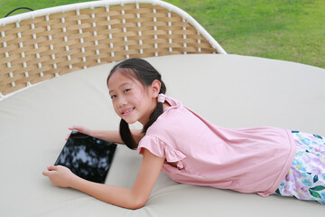 Smiling Asian girl child playing tablet and looking at camera while lying on fabric sofa in the garden. - 793795039