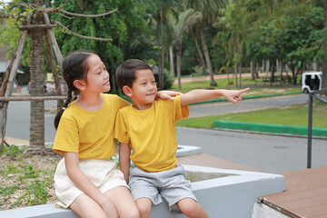Portrait of Asian boy and girl are sitting in the park and pointing at something beside them, Children are friends in the garden.