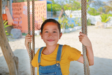 Poirtrait of Asian girl in dungarees jean with wind blowing hair on face sitting on swing at cafe near coast. - 793796267
