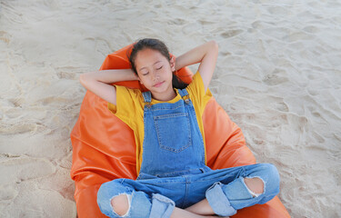 Smiling Asian girl child in dungarees jean relax and sleeping on orange sofa bed beach on sand at summer holiday.