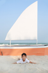 Happy Asian little boy looking straight at camera while lie down and play on sand against sailboat on the beach at summer holidays.