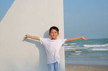 Portrait of smiling Asian boy open arms wide and looking at camera while standing on sailboat on the beach. - 793796863