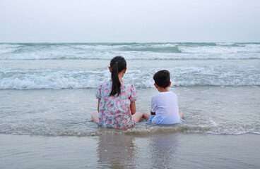 Rear of peaceful Asian young girl child and little boy sitting on tropical sand beach at sunset. Family of sister and brother in summer holiday. - 793797054