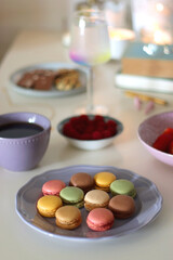 Obraz na płótnie Canvas Plate of pastel macarons, cookies and chocolate, cup of tea of coffee, glass of bubble water, various berries, books and accessories on the table. Selective focus, pastel colors.