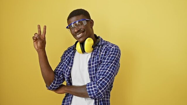 African american woman wearing safety glasses standing smiling with happy face winking at the camera doing victory sign. number two. over isolated yellow background