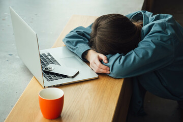 A tired woman lies face down in front of a laptop with a cup of tea.