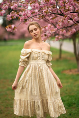 Obraz na płótnie Canvas Pretty young blonde girl in vintage lace dress standing in spring park near pink blossom flowers. Tenderness romantic model posing 
