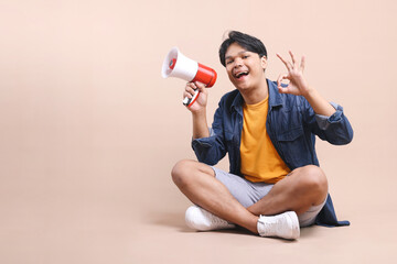 Full Body of Excited Young Asian Man Sitting and Announce Use Megaphone while Showing Ok Sign...