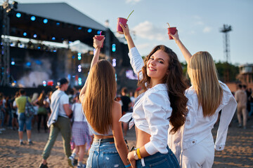 Girls with drinks enjoy live music at sunny beach festival. Group of friends dance, celebrate with...