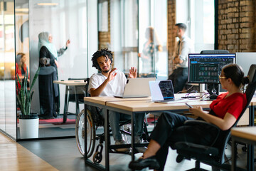 A young business group, including an African American businessman in a wheelchair, collaborates within a modern glass office, actively engaged around a computer and laptop, collectively solving