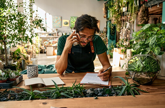 Successful confident business man plant shop owner working in greenhouse indoors. Florist talking on the phone, writing on a clipboard at a wooden work table with plants, decor and florarium.