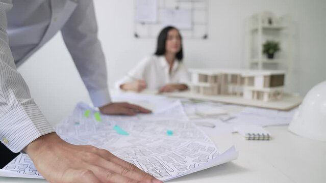 Skilled caucasian interior designer working with house model while discussing with asian architect engineer about house plan on meeting table with blueprint and house model scatter around. Immaculate.