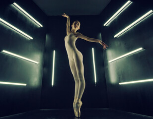 ballerina in a bodysuit and tights poses stretching in a dark photo studio in the light of lamps