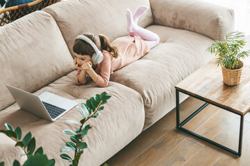 A cute 5-6-year-old girl wearing headphones while absorbed in her laptop screen, lounging...