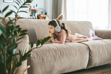 A cute little girl is absorbed in her laptop, wearing headphones to immerse herself in the digital...