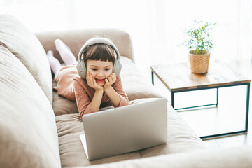 A charming 5-6-year-old girl lying on a sofa, wearing headphones and watching a laptop screen with focused attention